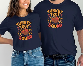 May include: Navy blue t-shirts with the text "TURKEY TROT SQUAD" in orange and red, featuring a cartoon turkey and autumn leaves. The shirts are worn by two people, suggesting a matching set for a Thanksgiving theme.
