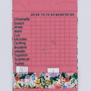 Peut inclure: Un organiseur d'aiguilles à coudre en tissu rose avec un motif floral. L'organisateur comporte une grille avec des numéros de taille d'aiguille et des étiquettes pour différents types de tissus. Plusieurs aiguilles à coudre sont visibles.
