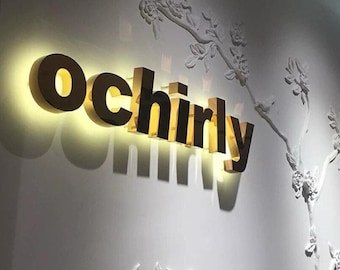 May include: A three-dimensional wall sign featuring the word ochirly in gold-toned, backlit lettering. The sign is mounted on a white wall decorated with a raised, white, sculptural floral branch relief that extends across the surface.