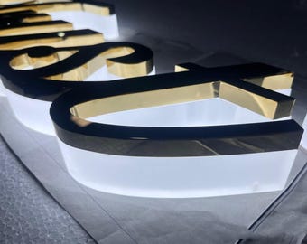 Könnte beinhalten: Ein dreidimensionales Schild mit goldfarbenen Metallbuchstaben in reflektierender Optik. Die Buchstaben sind auf einer durchscheinenden weißen Basis montiert, die ein sanftes Leuchten abgibt und einen Schichteffekt mit sichtbarer Tiefe erzeugt.