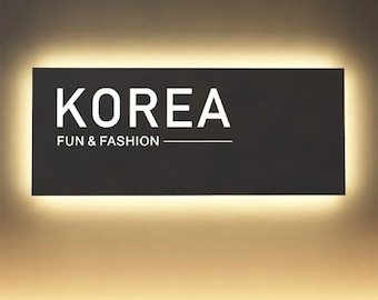 May include: A rectangular black wall sign with white text that reads KOREA FUN & FASHION. The sign features a bright, warm white LED backlight that creates a glowing halo effect around the edges of the dark, solid rectangular panel.