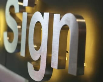 May include: A three-dimensional metal sign featuring the word Sign in large, brushed silver capital letters. The letters are mounted on a dark, flat surface and are illuminated from behind with a warm, golden glow that creates a halo effect.