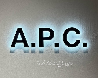 May include: Black illuminated sign spelling out "A.P.C." with a bright blue glow behind the letters. The sign is mounted on a light grey wall. Below the letters, the text "US Arts Desifn" is written in cursive.