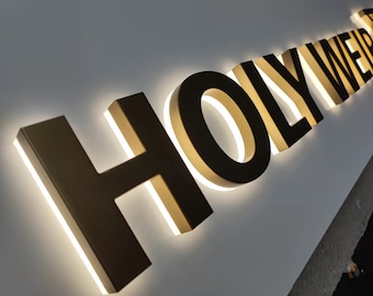 May include: A gold-coloured "HOLY WEIR" sign with white LED lights around the edges of each letter. The sign is mounted on a white wall.