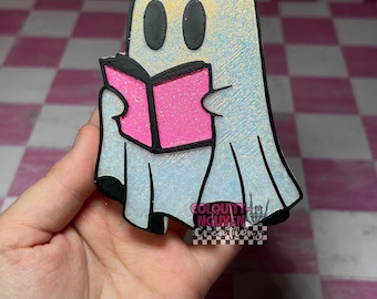 Op de afbeelding: Een 3D-geprint spookfiguur met een glinsterende iriserende witte afwerking, zwarte ovale ogen en een felroze open boek in de handen. De basis van het spookje is voorzien van een klein zwart-wit logo met de tekst Colquitt Nguyen Creations.