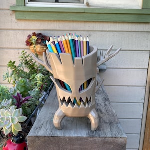 Può includere: Un portapenne beige a forma di albero dei cartoni animati con una faccia e corna, pieno di matite colorate. Il portapenne è su una superficie di legno, con una finestra e delle piante sullo sfondo.