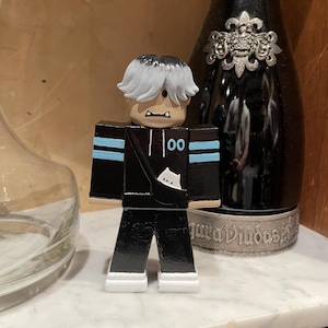 May include: A black and white figurine of a character with white hair and a black outfit with blue stripes. The figurine has a small bag with a cat design. The figure is standing in front of a black bottle.