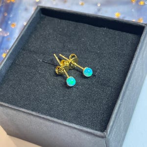 Puede incluir: Un par de pendientes dorados con pequeñas piedras redondas de color azul-verdoso opalino. Los pendientes se exhiben dentro de una caja de joyería negra, creando un contraste con los brillantes pendientes.