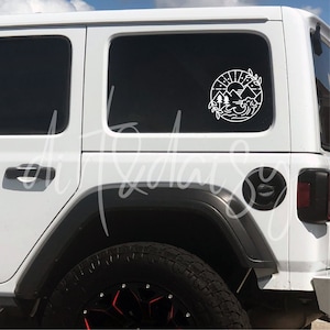 May include: White Jeep with a black and white sticker on the back window. The sticker is a circle with a mountain scene inside. The Jeep has a black tire with a red rim.