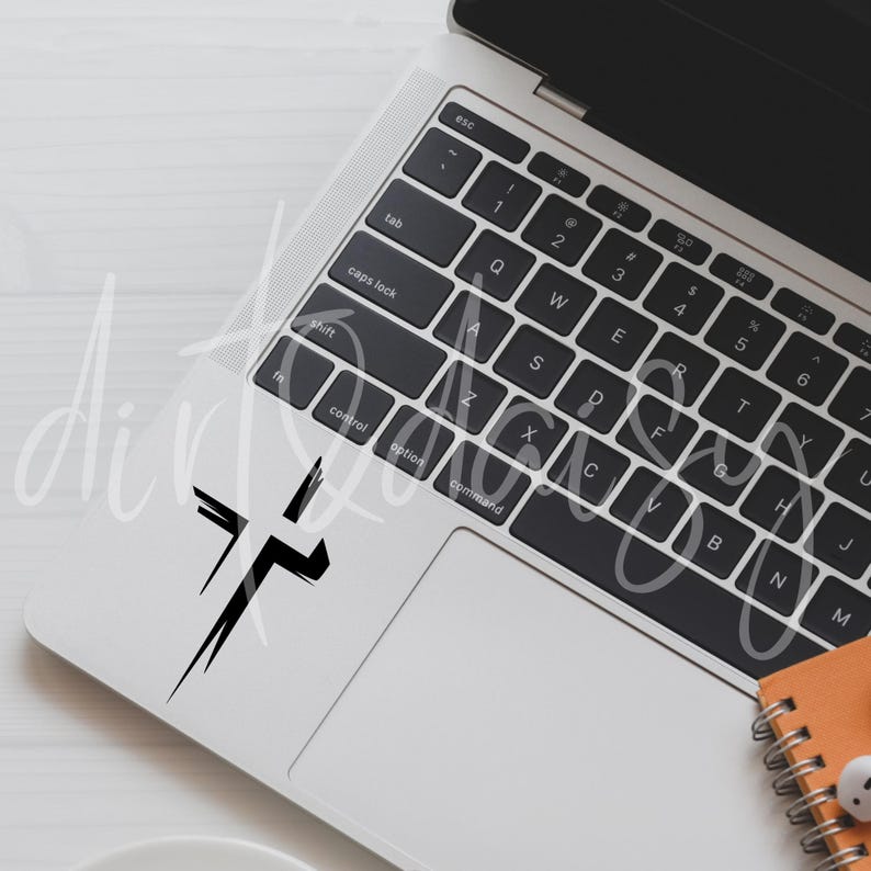 Pu&ograve; includere: Un adesivo a forma di croce nera &egrave; applicato nell'angolo in alto a sinistra di un laptop. Il design della croce &egrave; in stile pennellata. Il laptop &egrave; aperto, rivelando una tastiera e un trackpad. Un piccolo taccuino arancione e auricolari sono visibili.