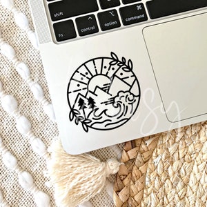 May include: Black and white sticker of a sun, mountains, and waves in a circle. The sticker is on a silver laptop.