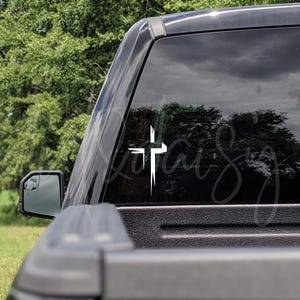 Pu&ograve; includere: Adesivo a forma di croce bianca sul lunotto posteriore di un camion nero. Il design della croce ha un aspetto invecchiato e strutturato con bordi affilati e frastagliati. Lo sfondo mostra una vista sfocata di alberi e uno specchietto laterale.