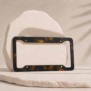 May include: A license plate frame with a tortoiseshell pattern. The frame is dark brown with yellow and black marbling. It has four mounting holes and a rectangular shape. The frame is displayed on a light-colored, textured surface.