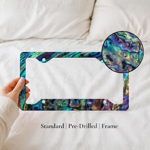 May include: A rectangular license plate frame with a vibrant, iridescent abalone shell design in shades of blue, green, and purple. The frame is pre-drilled and has the text "Standard | Pre-Drilled | Frame".