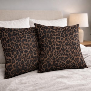 May include: Two square cushions featuring a brown and black leopard print design. They are placed on a bed with white bedding. These cushions add a touch of animal print to the bedroom decor.