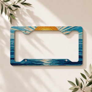 May include: A rectangular number plate frame featuring a mosaic-style design of a sunrise over the ocean. The top section displays a yellow and orange sun with radiating beams, while the lower portion depicts deep blue waves and water.