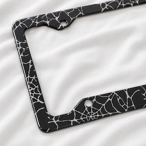Peut inclure: Un cadre de plaque d'immatriculation rectangulaire présentant une finition noire brillante avec un motif de toile d'araignée blanche imprimé sur la surface. Le cadre comprend deux trous de montage circulaires en haut et deux en bas.