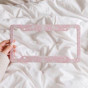 May include: A pink and white snakeskin-patterned license plate frame. The rectangular frame has rounded corners and four mounting holes. The frame is held by a hand against a white, wrinkled fabric background.