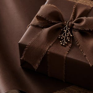 Könnte beinhalten: Eine quadratische Geschenkbox, eingewickelt in unifarbenes, dunkelbraunes Papier und gebunden mit einem breiten, dunkelbraunen Stoffband mit ausgefransten Kanten. Ein kleiner, metallischer, zweigförmiger Anhänger mit winzigen Perlen.