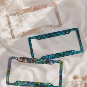 May include: Three decorative license plate frames with swirling patterns in various colours. One frame has a pink and white design, another is teal and blue, and the third features a mix of colours. Each frame has pre-drilled holes for easy installation.