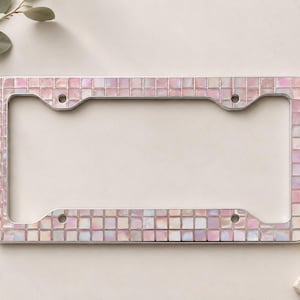 May include: A car number plate frame with a mosaic tile design in pink, white, and iridescent colours. The frame has a silver metal border and four mounting holes. A decorative accessory for a vehicle.
