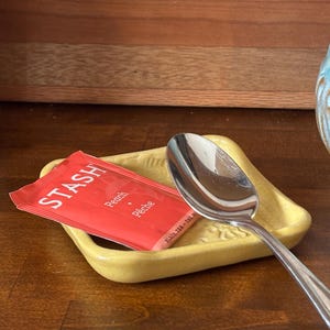 May include: A yellow ceramic spoon rest with a silver spoon and a red tea bag. The tea bag is labeled "STASH" with "Peach" and "Pêche" printed on it. The spoon rest has a decorative embossed design.