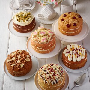 May include: Six different decorated cakes on white plates and cake stands. The cakes are topped with various toppings, including pistachios, almonds, white chocolate, caramel, and fruit.