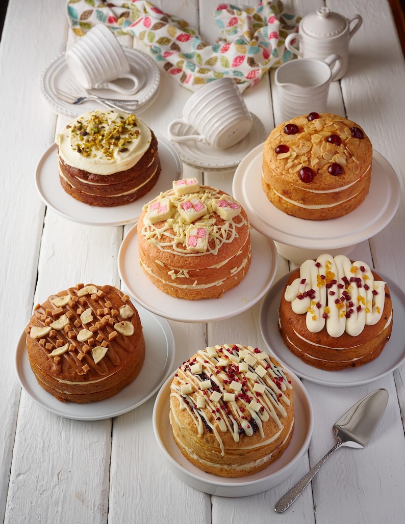 May include: Six different decorated cakes on white plates and cake stands. The cakes are topped with various toppings, including pistachios, almonds, white chocolate, caramel, and fruit.