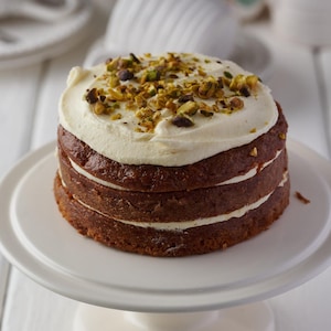 May include: A three-layer cake with white frosting and pistachio nuts on top. The cake is on a white cake stand.
