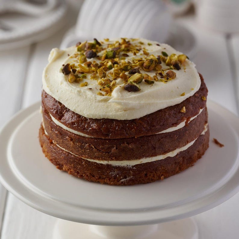 May include: A three-layer cake with white frosting and pistachio nuts on top. The cake is on a white cake stand.