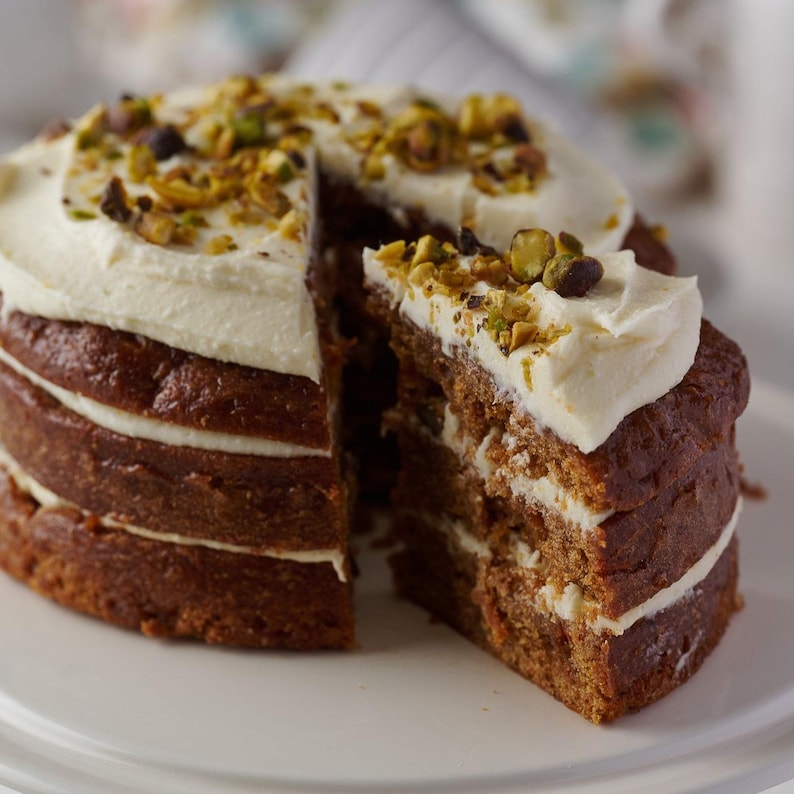 May include: A four-layer cake with white frosting and pistachio nuts on top. The cake is cut in half, revealing the layers of cake and frosting.