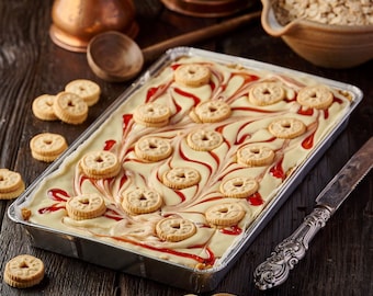 May include: A white chocolate and strawberry dessert topped with round cookies and a swirl of red strawberry sauce. The dessert is in a silver tray on a wooden table.