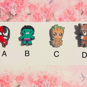May include: Four colorful cartoon character shoe charms, featuring Venom, Hulk, Groot, and Rocket Raccoon. The charms are arranged in a row on a white background.