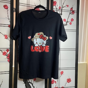 Puede incluir: Camiseta negra con un gr&aacute;fico blanco de Hello Kitty con un sombrero marr&oacute;n y sosteniendo un coraz&oacute;n rojo. El gr&aacute;fico dice "LOVE" en letras rojas.