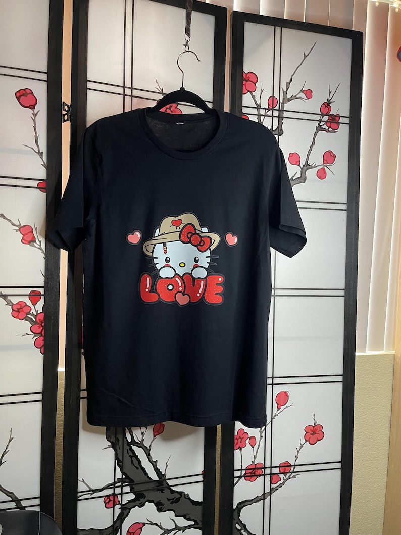 Puede incluir: Camiseta negra con un gr&aacute;fico blanco de Hello Kitty con un sombrero marr&oacute;n y sosteniendo un coraz&oacute;n rojo. El gr&aacute;fico dice "LOVE" en letras rojas.