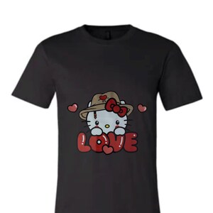 Puede incluir: Camiseta negra con una imagen de dibujos animados de Hello Kitty con un sombrero marr&oacute;n y un lazo rojo. El texto "LOVE" est&aacute; en letras rojas con corazones alrededor.