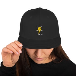 Puede incluir: Gorra de béisbol negra con un gráfico de rayo amarillo y la palabra "LIGHTNING" escrita alrededor.
