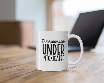 May include: White ceramic coffee mug with black text that reads "Overworked Under Intoxicated".