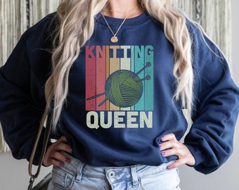 May include: A navy blue sweatshirt with a vintage-style graphic of a ball of yarn and knitting needles. The text "Knitting Queen" is printed below the graphic.