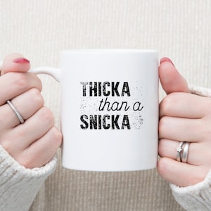 May include: White ceramic mug with black text that reads "THICKA than a SNICKA".