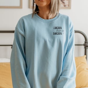 May include: A light blue sweatshirt with the text "Thicka than a Snicka" printed in black. The sweatshirt is a crew neck style and has long sleeves. The text is centered on the upper left chest.