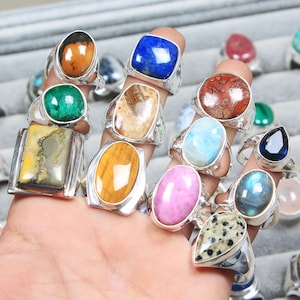 May include: A collection of gemstone rings with various shapes and colors, including blue lapis lazuli, green malachite, and tiger's eye. The rings have silver settings and are displayed on fingers.