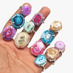 Puede incluir: Una colección de 10 anillos con rebanadas de ágata de colores engastadas en plata. Los anillos se muestran en una mano, mostrando los patrones y texturas únicos del ágata.