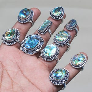 Może przedstawiać: Zestaw dziesięciu srebrnych pierścionków z wkładkami z muszli abalone. Pierścionki mają różne wzory, w tym owalne, w kształcie łzy i prostokątne. Muszla abalone prezentuje tęczowe kolory niebieskiego, zielonego i złotego.