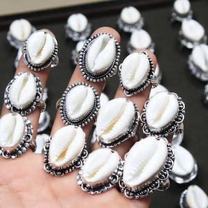 Op de afbeelding: Een verzameling zilveren ringen met witte kauri-schelpen. De ringen worden op een hand getoond, waardoor de ingewikkelde details van de schelpontwerpen worden getoond.