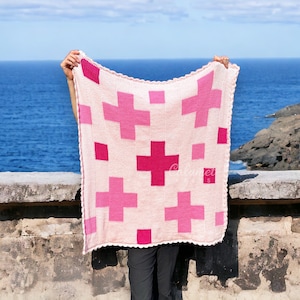 May include: A pink and white knitted blanket with a plus sign pattern. The blanket is being held up in front of a blue ocean and rocky coastline.