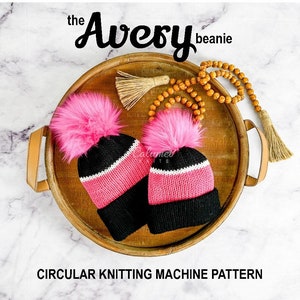May include: Two pink and black knit beanies with fluffy pink pom poms on a wooden tray. The beanies are labelled "the Avery beanie" and "Circular Knitting Machine Pattern".