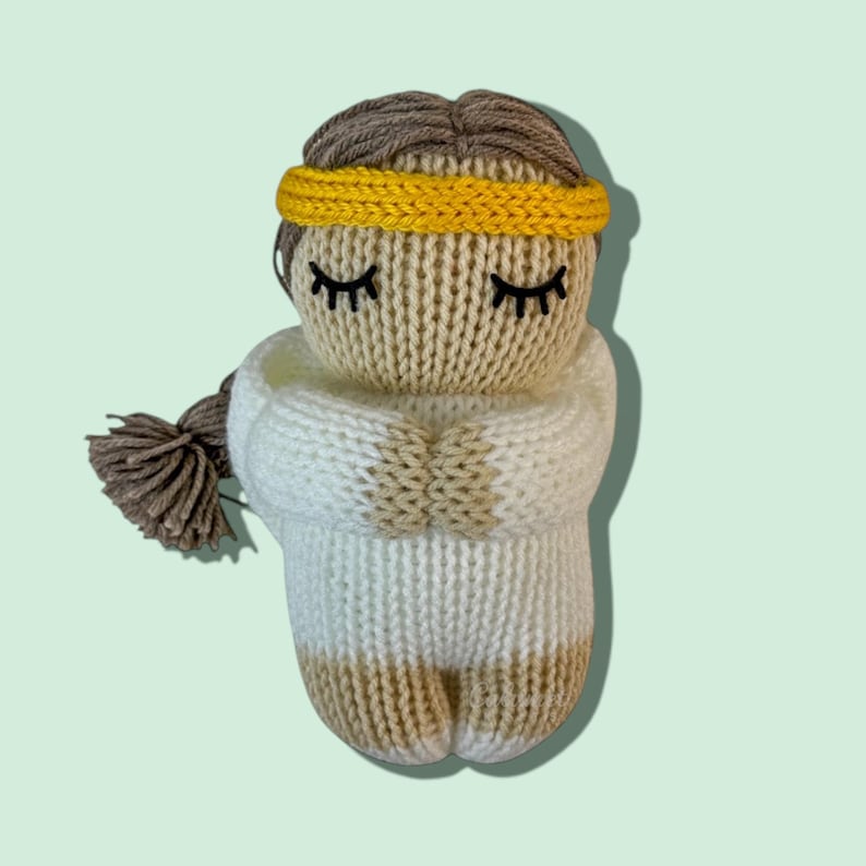 K&ouml;nnte beinhalten: Eine gestrickte Puppe mit geschlossenen Augen, braunen Haaren und einem gelben Stirnband. Die Puppe tr&auml;gt ein wei&szlig;es und beiges Strickoutfit.