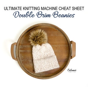 May include: A white knitted beanie with a brown faux fur pom pom on top, sitting on a wooden tray. The beanie is a double brim style. The text "Calumet Knits" is visible in the lower right corner of the image.