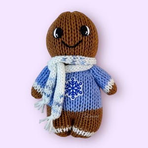 K&ouml;nnte beinhalten: Eine braune gestrickte Lebkuchenfigur, die einen blauen Pullover mit einem wei&szlig;en Schneeflockenmuster und einen wei&szlig;-blau gestreiften Schal tr&auml;gt.